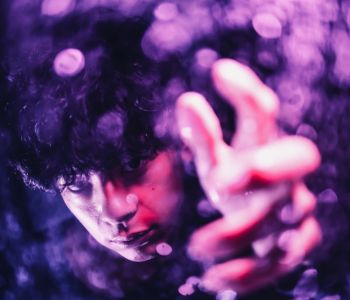 紫の照明の中、レンズに手を伸ばす男性アーティストを幻想的に捉えたポートレート。