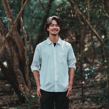 柔らかな笑顔の男性が緑の木々に囲まれた森の中で立つポートレート写真